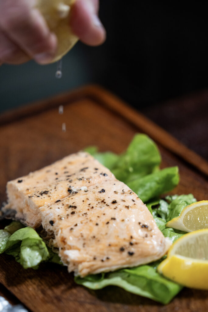 Close-up of a hand squeezing a fresh lemon over a piece of grilled salmon served on a bed of greens.