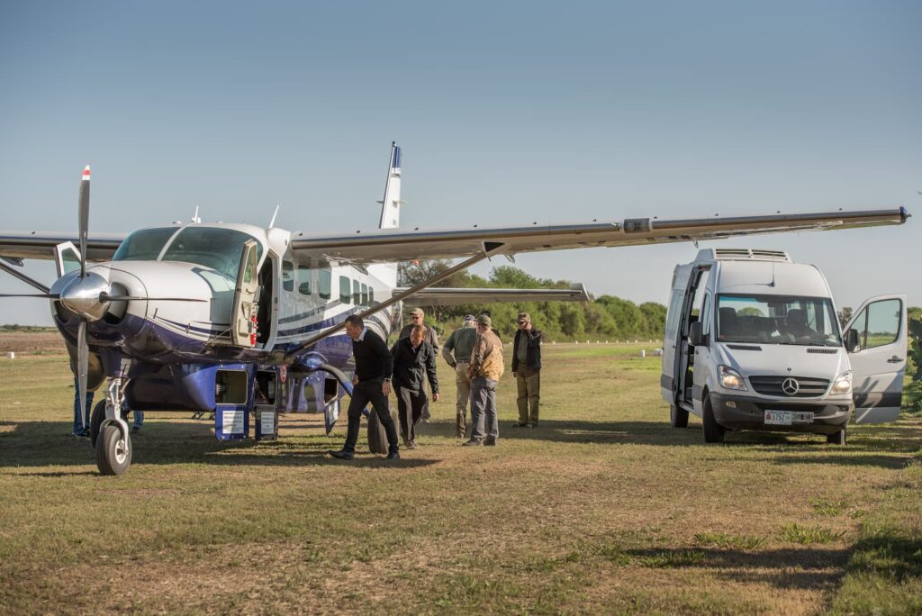Hunters with their gear beside a single-engine charter aircraft and a transfer van on a grass airstrip