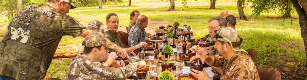 Hunters share an asado lunch at a long table under the trees while an H&H staff member serves the group